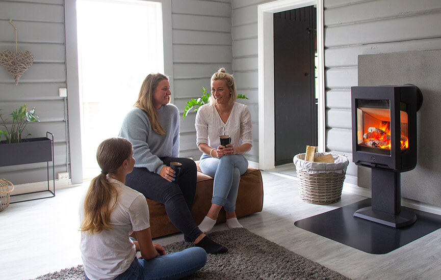 Tre piger sidder på en brun puf i en grå stue foran Jøtul F 167 brændeovnen